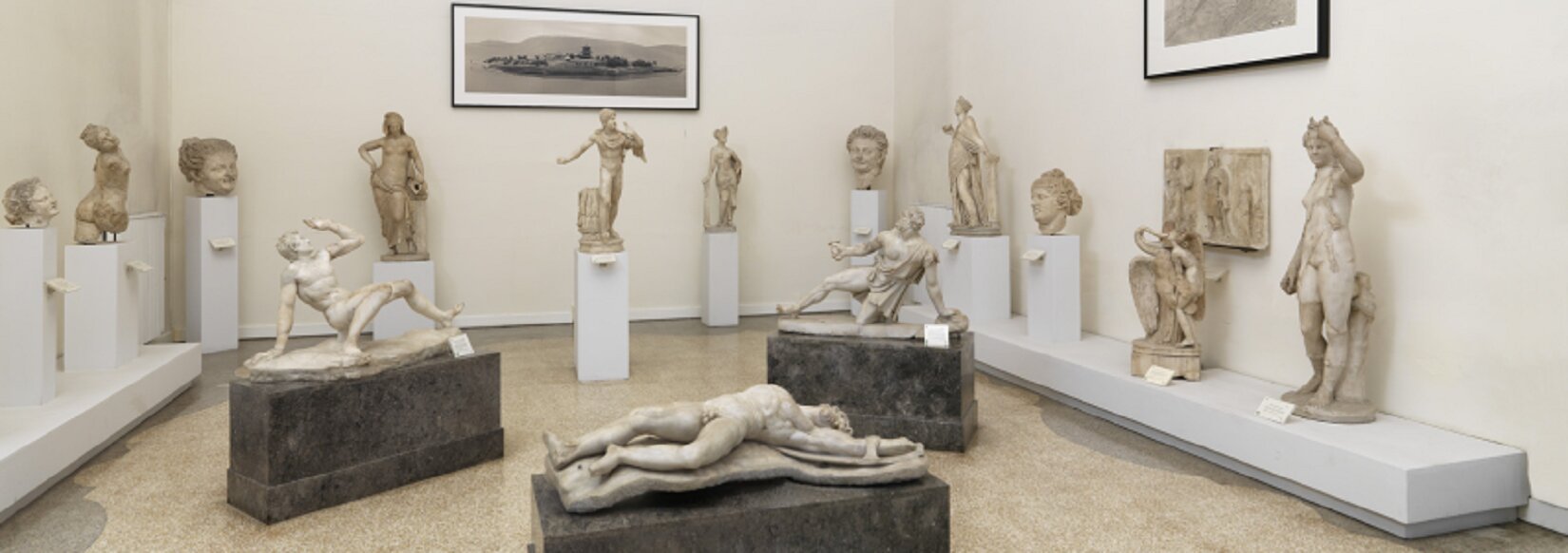 Ilustracja przedstawia fotografię „Rzeźby rzymskie w Narodowym Muzeum Archeologicznym w Neapolu”. Na zdjęciu znajduje się pomieszczenie o białych ścianach i beżowej podłodze. Na środku pomieszczenia są trzy grafitowe podesty, na których są rzeźby. Rzeźby wykonane się z jasnego kamienia i przedstawiają mężczyzn. Pierwszy mężczyzna leży na plechach, drugi kuca i opiera się prawą dłonią, a trzeci klęczy na lewym kolanie. Wzdłuż ścian stoją białe podesty, na których są mniejsze rzeźby wykonane z jasnego kamienia.  Przedstawiają postacie ludzkie lub same głowy. Na ścianach wiszą dwa biało‑czarne obrazy w czarnych ramkach i jedna płaskorzeźba z jasnego kamienia. 