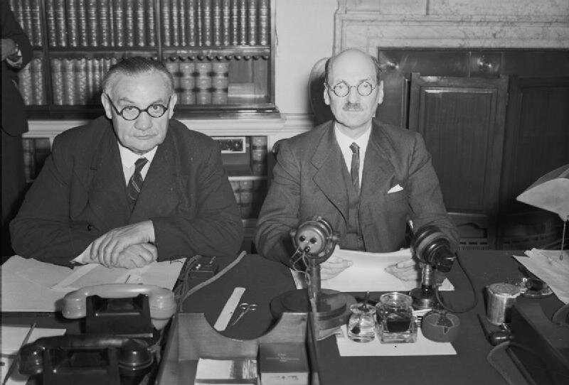 Zdjęcie przedstawia dwóch starszych mężczyzn w garniturach siedzących przy biurku. Na biurku leżą rozłożone papiery, stoją dwa telefony i dwa mikrofony.
