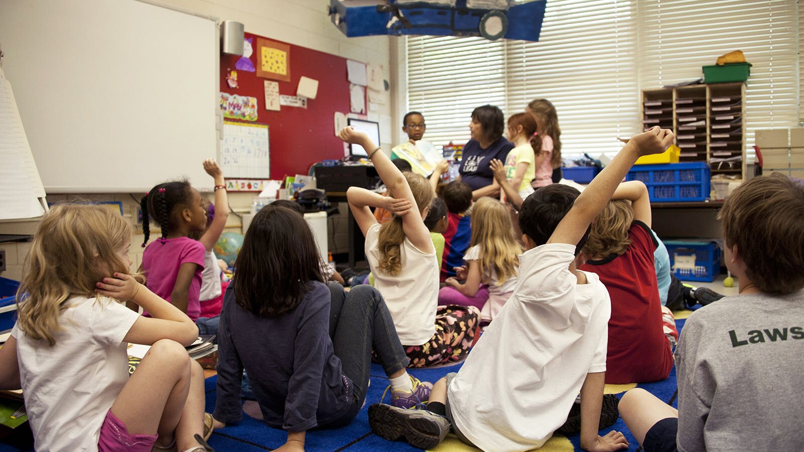 Zdjęcie przedstawia grupę kilkunastu dzieci siedzących na podłodze, przed kobietą siedzącą przy oknach, obok której stoi troje dzieci, chłopiec i dwie dziewczynki. Ujęcie zostało zrobione zza pleców dzieci siedzących na podłodze.