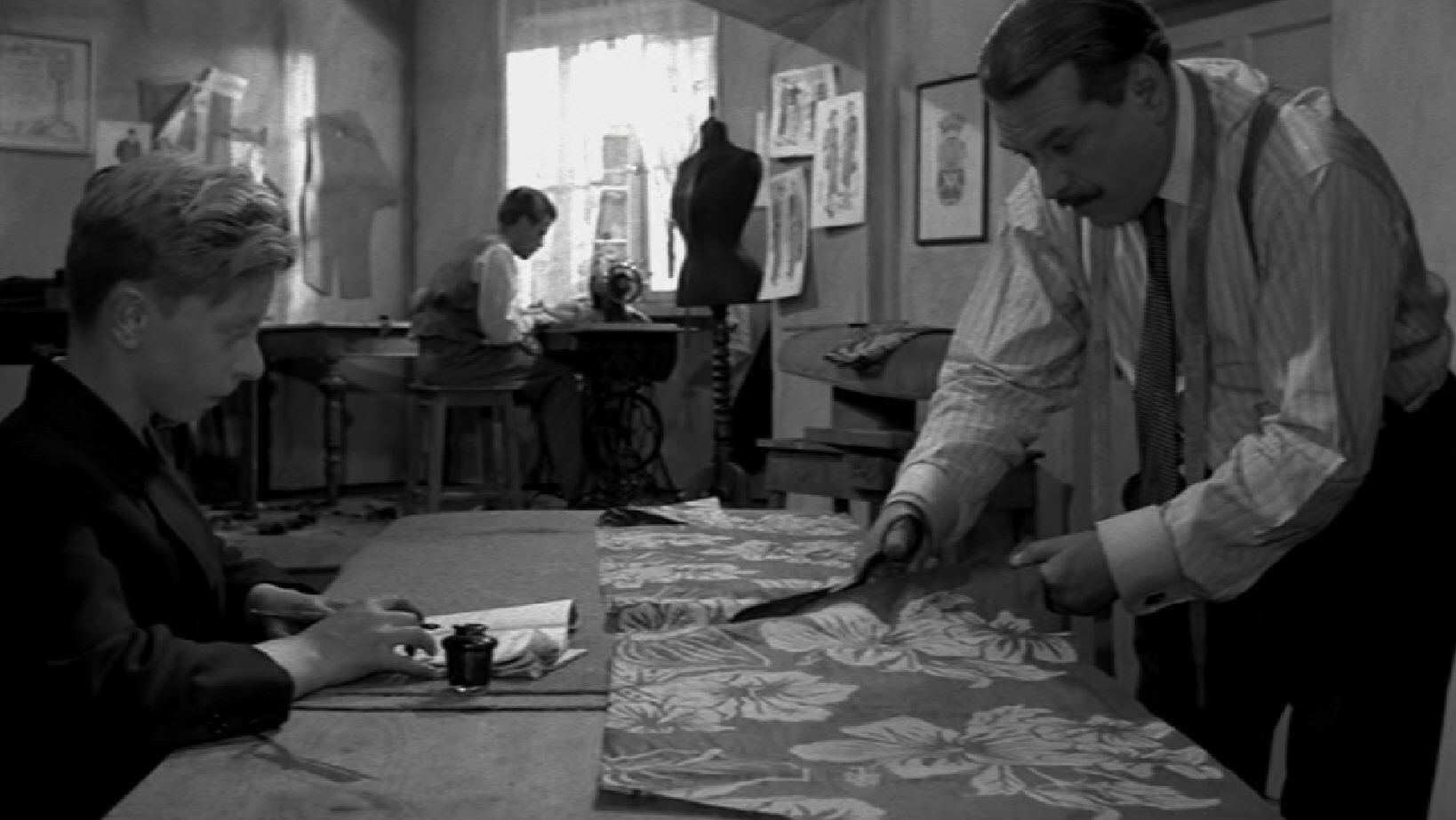 Zdjęcie przedstawia chłopca siedzącego przy stole, odrabiającego lekcje. Przy tym samym stole dorosły mężczyzna tnie nożyczkami wzorzysty materiał. Na drugim planie znajduje się drugi mężczyzna, szyjący na maszynie.