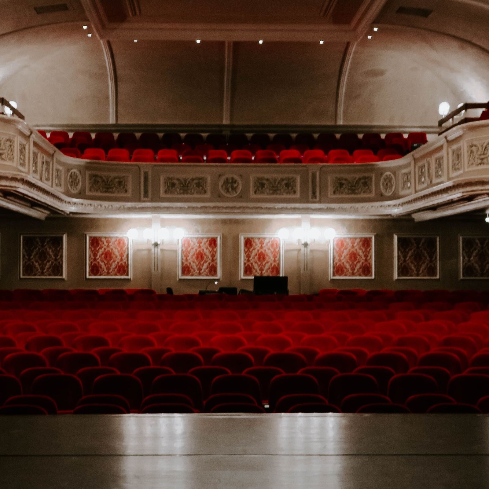 Zdjęcie przedstawia salę w teatrze od strony sceny. Widoczne są krzesła wyściełane czerwonym suknem, balkony z rzędem identycznych krzeseł oraz ściany, na których wiszą grafiki.  
