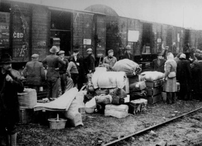 Zdjęcie przedstawia bocznicę kolejową, przy której stoi pociąg z szeregiem towarowych wagonów. Przy wagonach stoją ludzie ze swoim dobytkiem: ubraniami, pościelą, miskami, garnkami, koszami, walizkami.