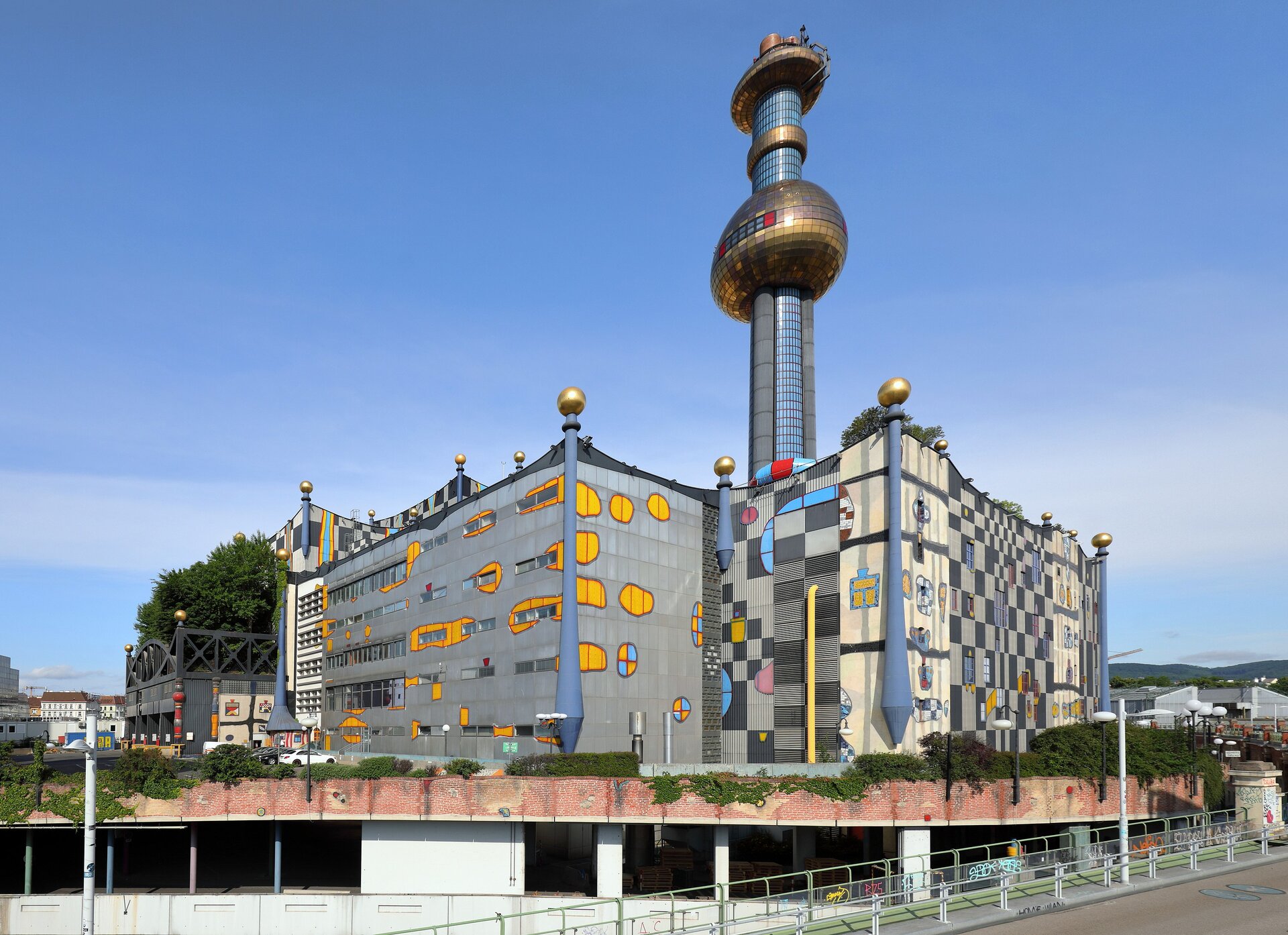 Zdjęcie przedstawia spalarnię Spittelau w Wiedniu. Budynek ma futurystyczny kształt. W rogach prostokątnego budynku znajdują się szare kolumny rozszerzające się do dołu, zwieńczone u góry złotymi kulami. Na fasadzie w kolorze szarym znajdują się nieregularne, pomarańczowe i niebieskie plamy oraz inne fantazyjne kształty oraz szaro – czarna szachownica. Nad budynkiem góruje srebrna wieża ze złotą kulą w połowie wysokości, złotą obręczą w 3/4 wysokości oraz płaską platformą na szczycie. 