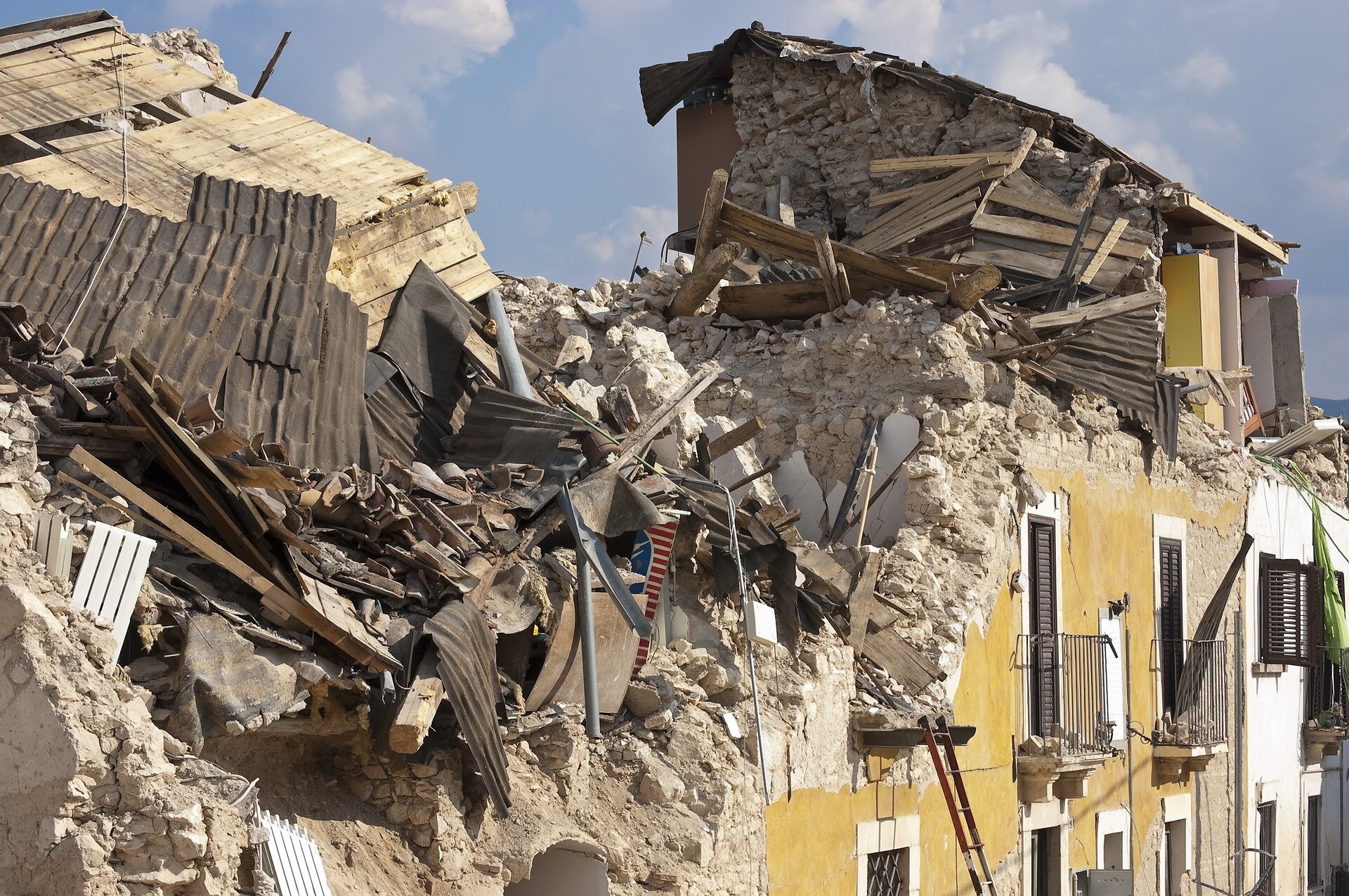 Zdjęcie przedstawia ruiny budynku zniszczonego w wyniku trzęsienia ziemi we Włoszech. Ściany domu zawaliły się do środka. Widoczny jest gruz, pozostałości dachu, mebli. Część budynku ocalała i stoi niezniszczona. Budynek jednak nie nadaje się do zamieszkania.