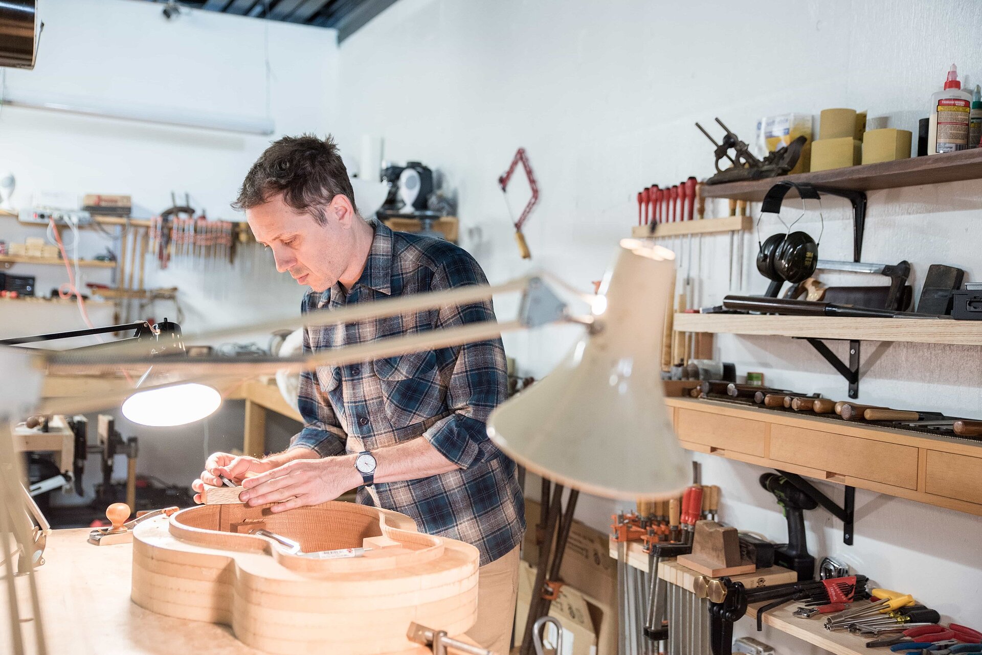 Ilustracja przedstawia fotografię warsztatu lutnika. Lutnik stoi przy stole, na którym leży niedokończony instrument. Mężczyzna przymocowuje do niego drewniany element. Ma na sobie koszulę w kratę i jasne spodnie. Na stole stoją dwie zapalone lampy oraz są narzędzia. Za mężczyzną, na ścianie, znajdują się półki pełne różnorodnych narzędzi, klejów, taśm, słuchawki, imadła, śrubokręty i tym podobne. Ściana w warsztacie jest biała i w większości zapełniona.