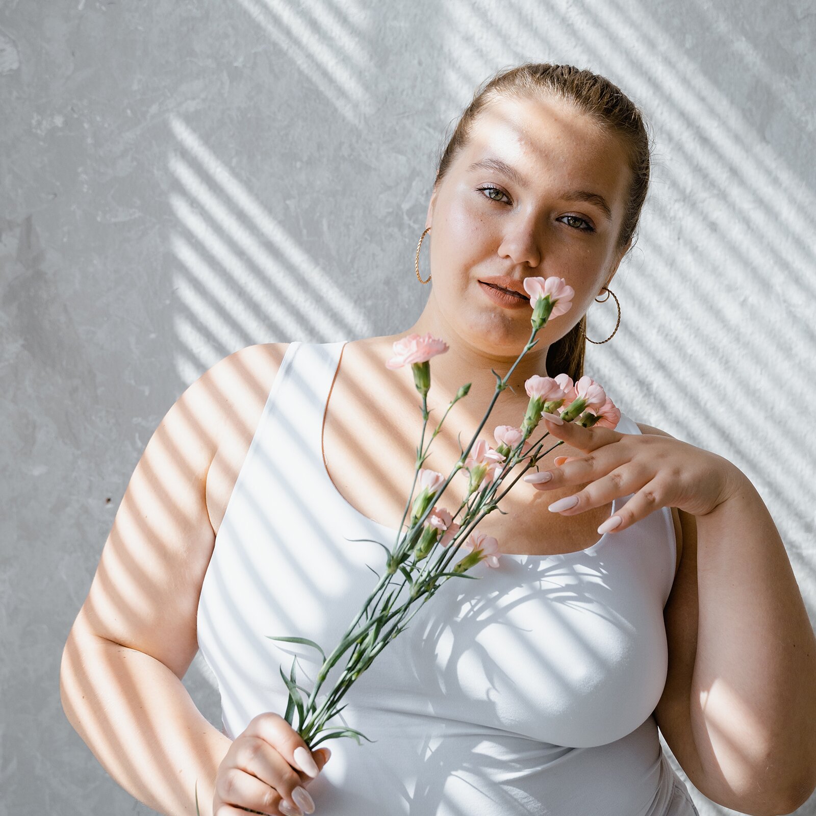 Zdjęcie przedstawia ładną, wysoką i grubszą kobietę. Ma ona długie, proste blond włosy zebrane w kucyk. Ma jasne oczy. Jest ubrana w biały podkoszulek, a w ręce trzyma kwiaty.