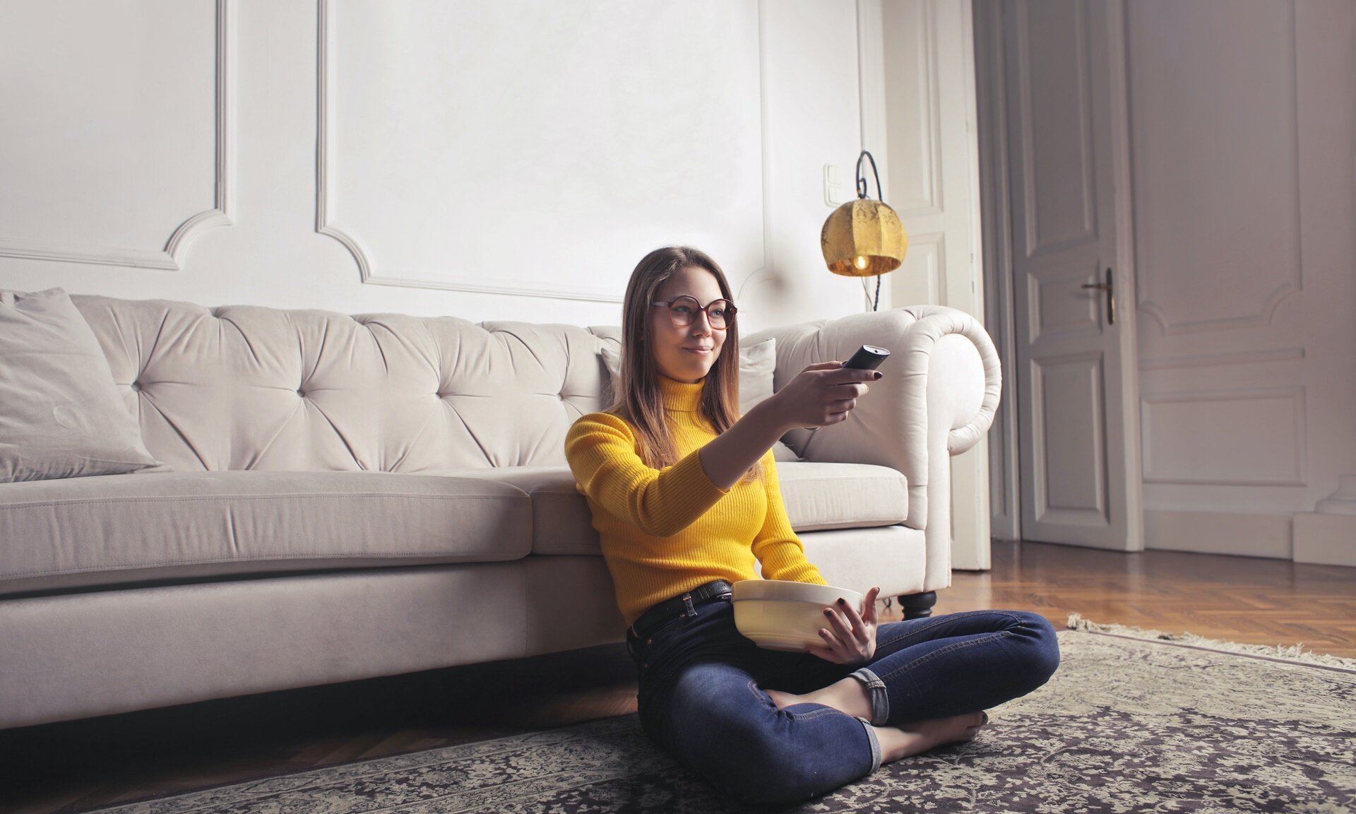 Zdjęcie przedstawia młodą kobietę, która siedzi na dywanie w salonie. Kobieta ma długie rozpuszczone włosy, okulary i jest ubrana w żółty golf oraz dżinsowe spodnie. Jej nogi są skrzyżowane, plecami opiera się o dużą, beżową sofę. W prawej ręce wyciągniętej do przodu i lekko uniesionej trzyma pilot, zaś lewą ręką podtrzymuje miskę, która oparta jest o jej brzuch. W tle widoczne są białe ściany pomieszczenia, otwarte białe drzwi oraz lampa.