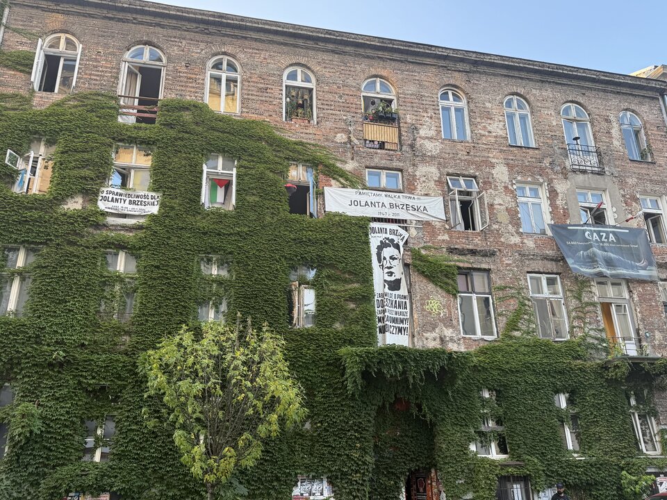 Kamienica mieszkalna w Warszawie objęta procesami reprywatyzacyjnymi. Ceglany budynek o widocznych śladach zużycia i nierównej powierzchni ścian, częściowo porośnięty zielonym bluszczem. Na ścianach umieszczono banery upamiętniające Jolantę Brzeską - lokatorkę i działaczkę społeczną zaangażowaną w obronę praw mieszkańców kamienic oraz banery odnoszące się do konfliktu izraelsko‑palestyńskiego.
