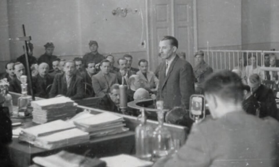 Zdjęcie przedstawia ludzi zgromadzonych w sali sądowej podczas procesu. W centralnym punkcie, w miejscu wydzielonym dla świadka,  stoi mężczyzna w średnim wieku, który składa zeznania. Ubrany jest w marynarkę, spodnie, koszulę i krawat. Widzimy jego lewy profil. Ma szczupłą twarz i krótkie włosy zaczesane do góry.  Naprzeciwko niego stoi duży stół na którym leżą dokumenty oraz szklane karafki na wodę. Za stołem siedzi skład sędziowski. Obok widzimy oparty o stół krzyż. Po lewej stronie zdjęcia, na ławkach, w eskorcie żołnierzy, siedzi kilkunastu mężczyzn. Przed nimi poniżej siedzą ich obrońcy.  Na sali zgromadzeni są jeszcze przedstawiciele prasy, na co może wskazywać mikrofon z boku stołu sędziowskiego oraz ludzie obserwujący przebieg procesu, którzy siedzą za barierką.  