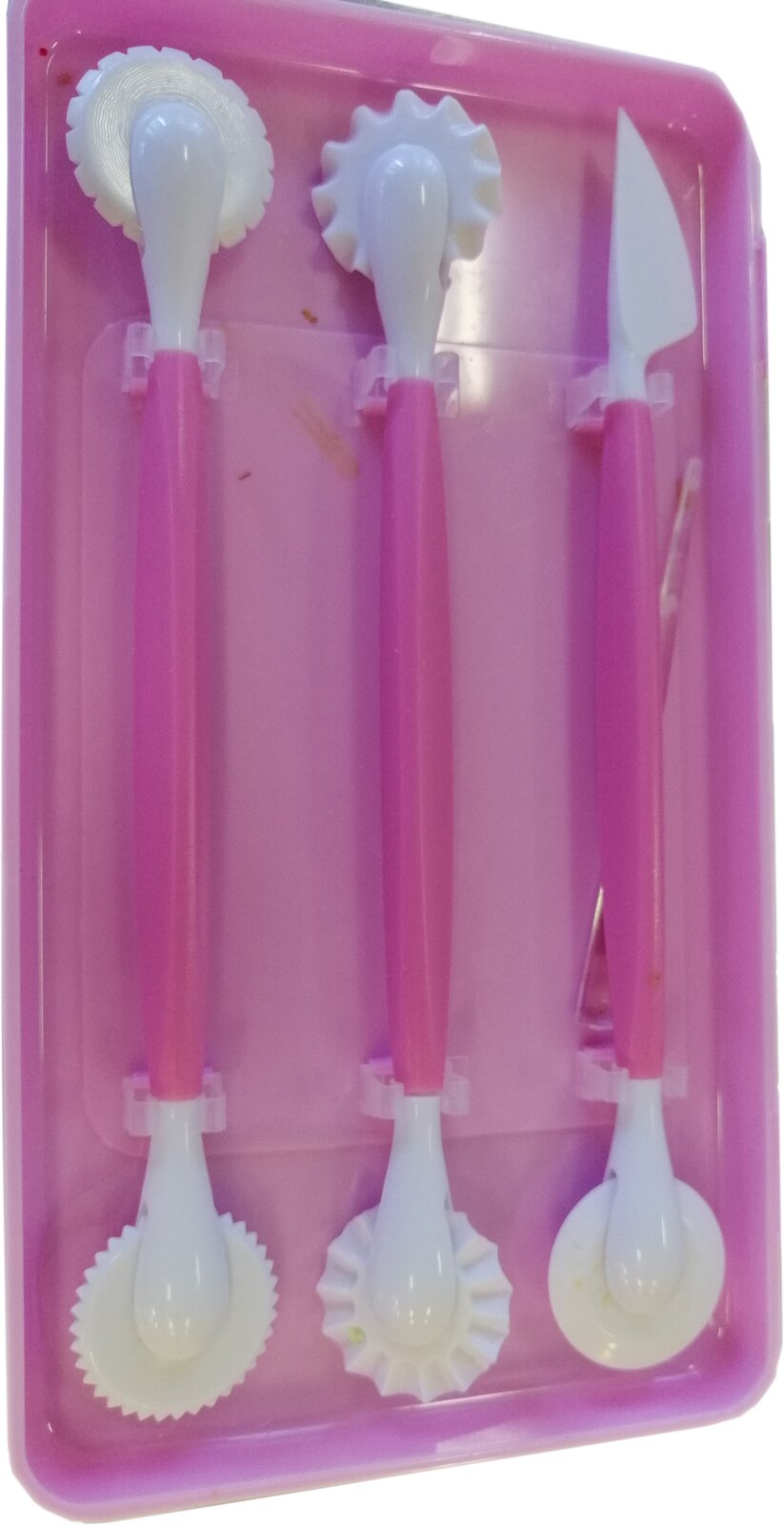 Zdjęcie przedstawia radełka. Widoczne są trzy radełka ułożone obok siebie w różowym etui. Każde radełko składa się z cylindrycznego korpusu w kolorze różowym, który jest również rękojeścią. Na obu końcach rękojeści znajdują się białe części robocze radełka w kształcie gładkich tarcz, ząbkowanych tarcz i nożyka.