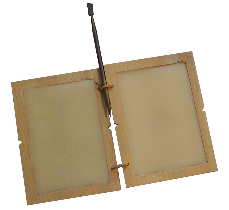 Zdjęcie przedstawia dwie połączone ze sobą tabliczki za pomocą dwóch metalowych kółeczek. Karty obramowane jasną, drewnianą ramą. Pomiędzy górne kółeczko i dyptyk włożono metalowy szpikulec- stylos.