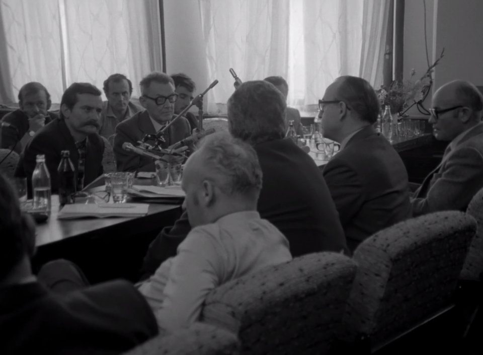 Ilustracja przedstawia mężczyzn siedzących za stołem, rozmawiających ze sobą. Mają rozłożone papiery, dokumenty. Są ubrani w garnitury, mają skupione twarze. Za nimi siedzą inni ludzie. Ustawione są mikrofony. 