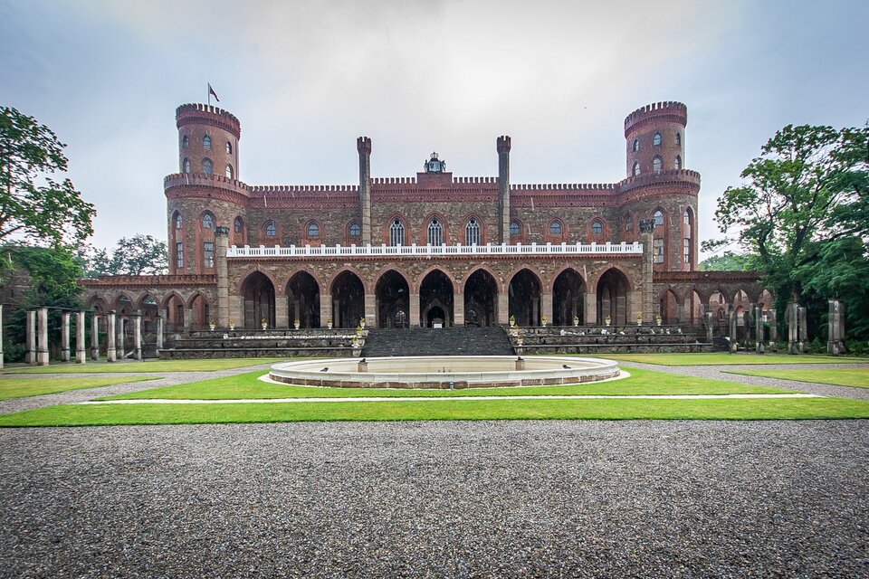 Fotografia przedstawia budynek pałacu. Na pierwszym planie znajduje się wjazd do budynku. Przed wejściem umieszczono okrągłą fontannę, która nie jest czynna. Do pałacu wiodą szerokie schody. Przed wejściem znajdują się ozdobione kolumnami krużganki. Nad krużgankami rozciąga się obszerny taras. Okna są wysokie i wąskie. Po bokach znajdują się po baszty. Dolna część jest większa, górna mniejsza. Na szczycie lewej baszty umieszczony jest maszt, na którym powiewa flaga. Po bokach budynku rosną drzewa. Niebo w tle jest lekko zachmurzone.