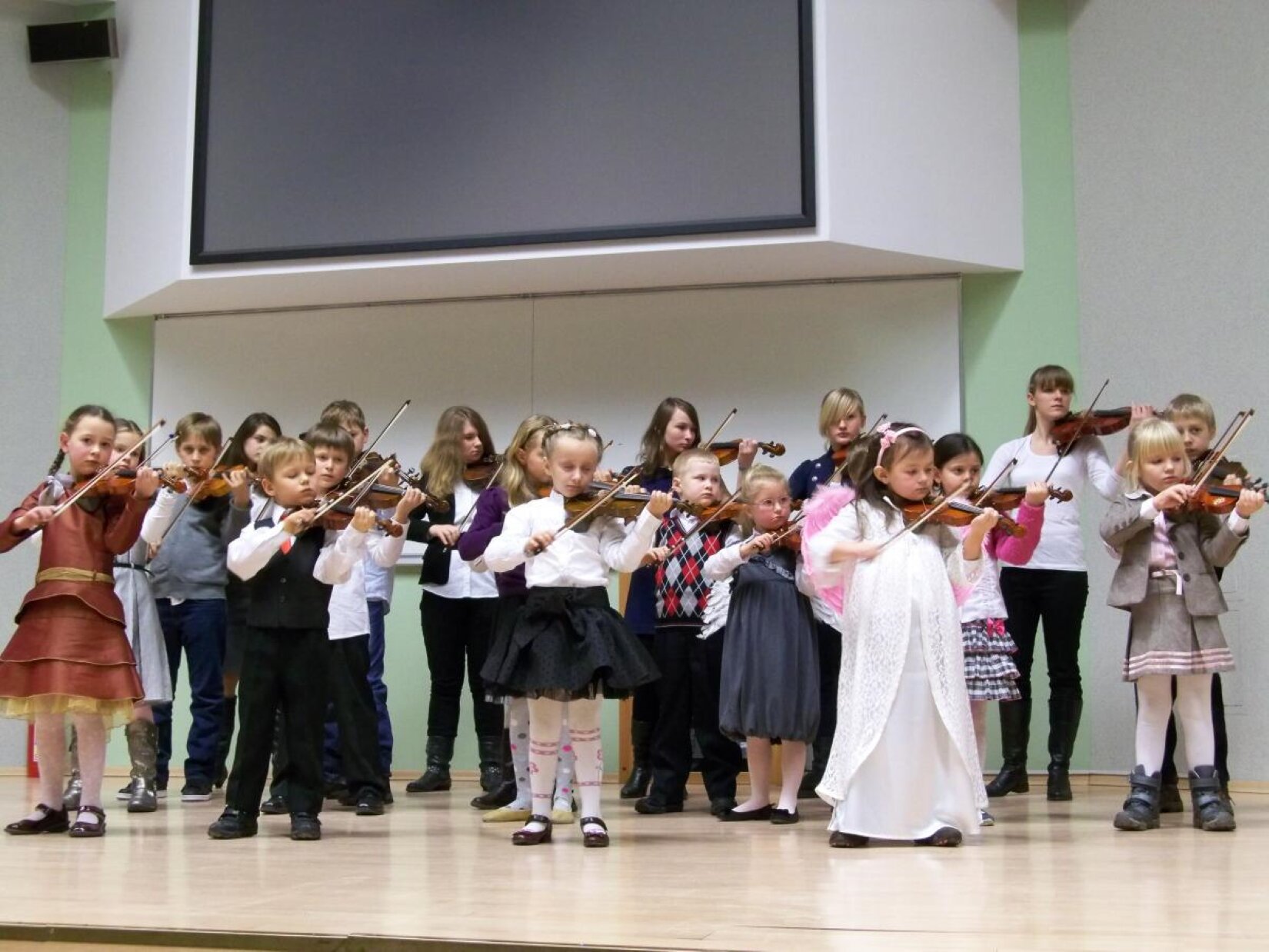 Fotografia z koncertu dziecięcego z Częstochowskiej Szkoły Suzuki, przedstawia na scenie sporą grupkę dzieci w wieku szkoły podstawowej, które odgrywają koncert muzyczny do jasełek. Każde dziecko trzyma prawidłowo ułożone dłonie do grania na skrzypcach. Wszystkie dzieci ubrane są na galowo, w tym jedna dziewczynka stojąca na pierwszym planie po prawej stronie jest przebrana za aniołka w białej, długiej sukience, różowymi skrzydłami na plecach oraz opaską z małą kokardką po prawej stronie głowy, która jest idealnie dobrana pod kolor skrzydeł. Oświetlenie na Sali jest bardzo wyraziste. Sala ma biało‑zielone ściany. W tle wisi duży, plazmowy telewizor w czarnym kolorze. 