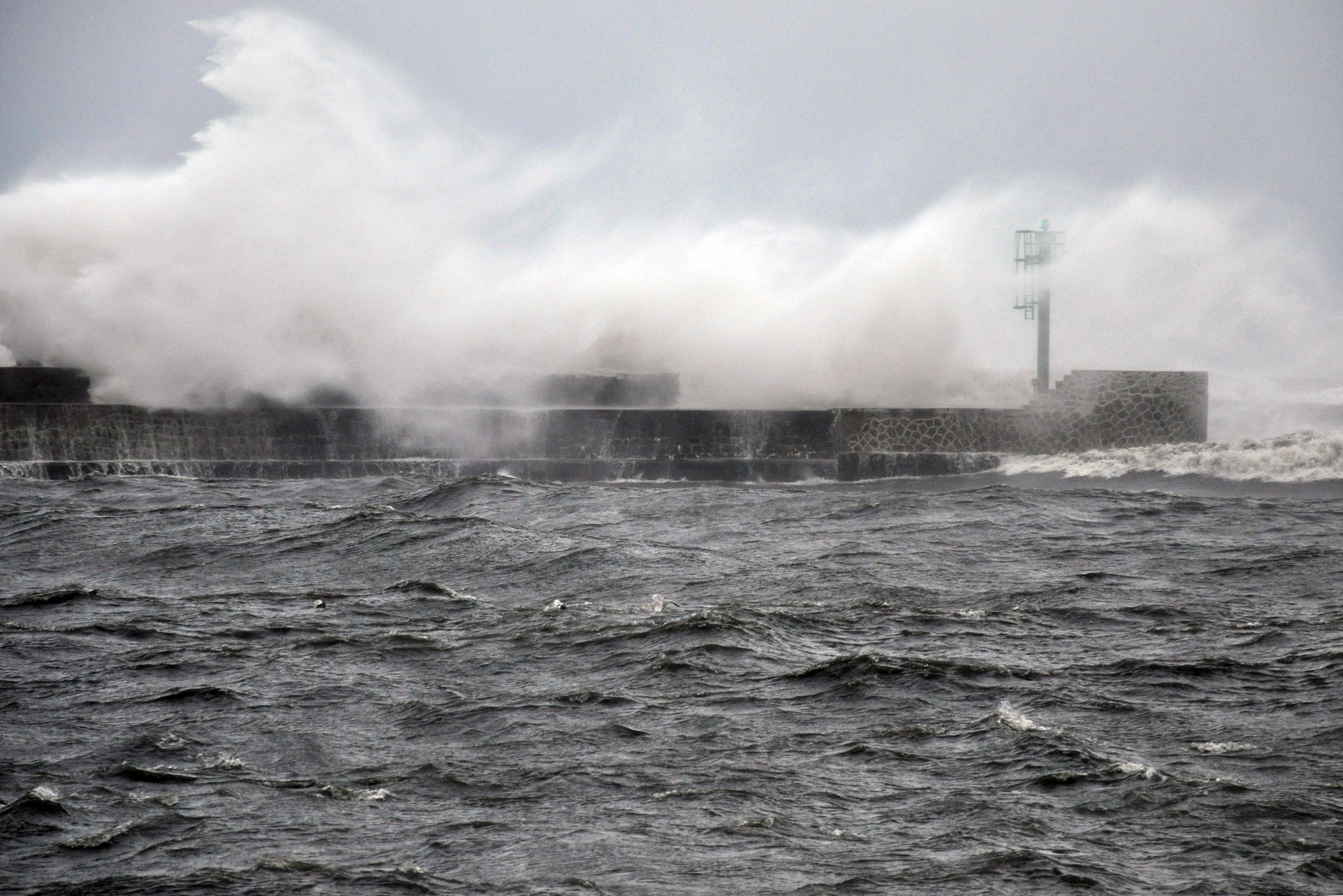 Fotografia prezentuje wygląd wybrzeża morskiego podczas huraganu. Widoczne wzburzone morze oraz fale rozbryzgujące się o molo.