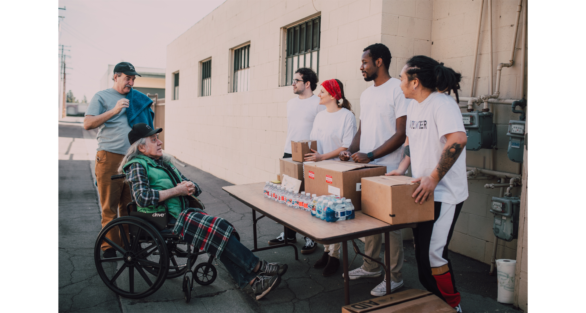 Zdjęcie przedstawia czworo  wolontariuszy. Stoją przy stoliku, na którym znajdują się kartonowe pudełka oraz butelki z wodą. Przed stolikiem znajduje się mężczyzna na wózku inwalidzkim. Obok niego stoi inny mężczyzna.