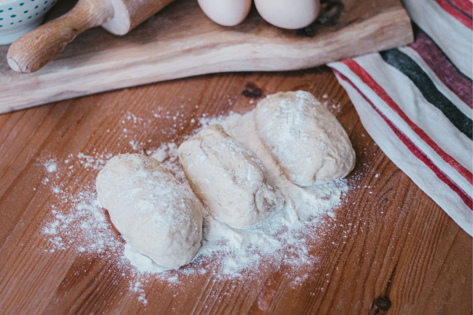 Zdjęcie przedstawia kontynuację kroku pierwszego formowania wianka debrzeńskiego. Ujęcie wykonane jest od góry pod skosem. Widoczny jest ciemnobrązowy blat na którym w środkowej części zdjęcia znajdują się trzy owalne kęsy ciasta oprószone białą mąką. Na blacie dookoła ciasta rozsypana jest mąka. W górnym lewym rogu grafiki dostrzec można fragment białej miseczki w niebieskie małe kropki, a obok niej widoczna jest drewniana jasnobrązowa rączka wałka do ciasta. Po prawo od wałka znajdują się dwa jajka. Wszystkie elementy leżą na drewnianej jasnobrązowej desce. W prawej części zdjęcia widoczna jest biała ścierka w czarne i czerwone pasy ułożona pionowo. Róg ścierki znajduje się pod drewnianą deską.