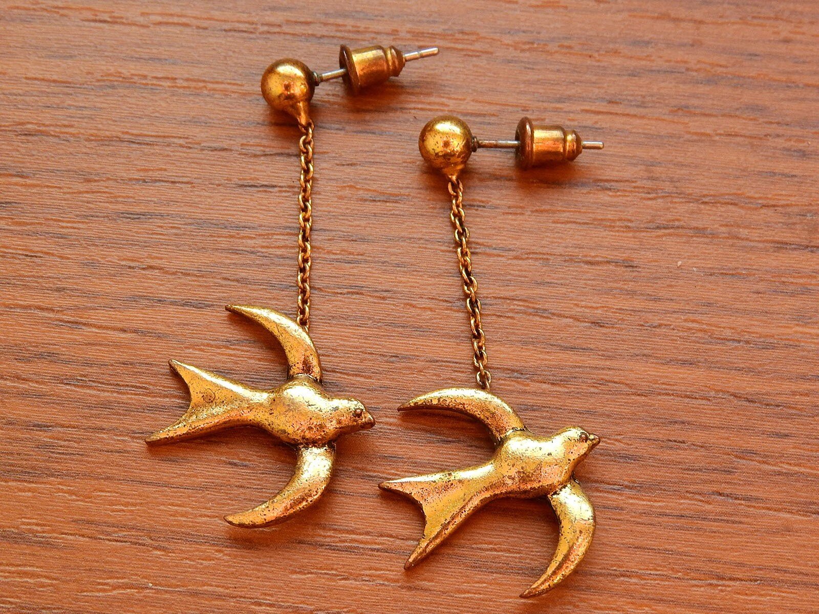 Zdjęcie przedstawia parę złotych kolczyków w kształcie ptaków, leżących na drewnianym blacie. Kolczyki są zapinane na sztyft, zakończony cylindrycznym zaciskiem. Figurki ptaków, wiszące na krótkim łańcuszku, mają rozpostarte do lotu skrzydła.