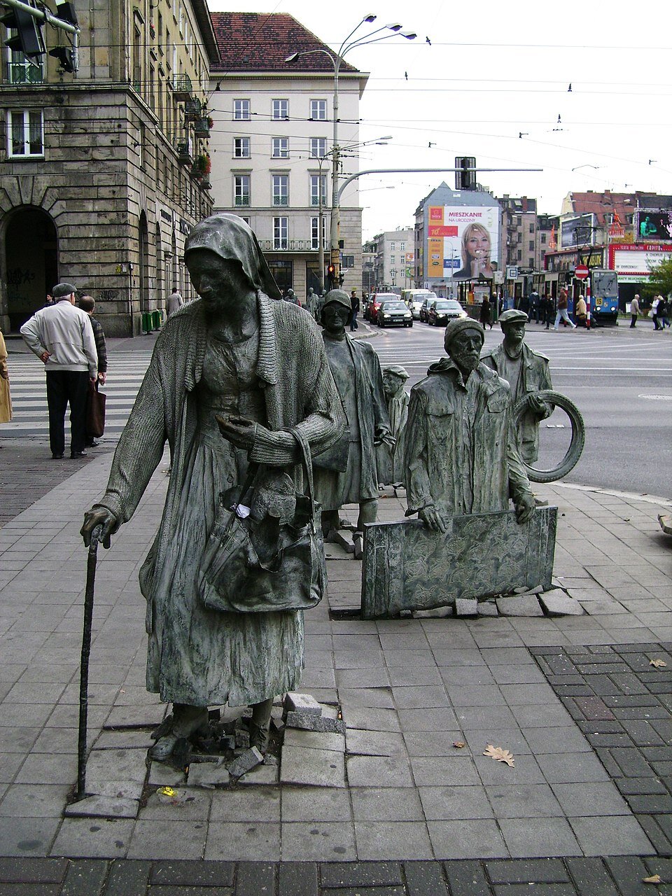Zdjęcie przedstawia postaci odlane z brązu umieszczone w miejskiej przestrzeni - ulicznym chodniku. Postacie „wynurzają się” z chodnika, jakby wychodziły z podziemnego przejścia. Przypominają one zastygłych w bezruchu przechodniów. Osoba na pierwszym planie to starsza kobieta. Ma opuszczoną głowę, na głowie chustkę, ubrana jest w wyciągnięty sweter, podpiera się laską, w drugiej ręce trzyma starą zniszczoną dużą torbę. Za nią znajduje się kolejna kobieta w kapeluszu z torbą trzymaną w prawej dłoni. Po prawej stronie kobiety postawiono odlaną z brązu figurę mężczyzny, trzyma on w dłoniach dużą płytę. Za nim kolejna postać — mężczyzna z oponą w dłoniach, następna osoba ukazana jest tylko do połowy, wygląda, jakby zapadła się w głąb ulicznego chodnika. Im bliżej posągi osób znajdują się ulicznej krawędzi widocznej w tle, tym głębiej "zanurzone są" w chodnikowych płytach. W tle ulica miasta z samochodami i budynkami.