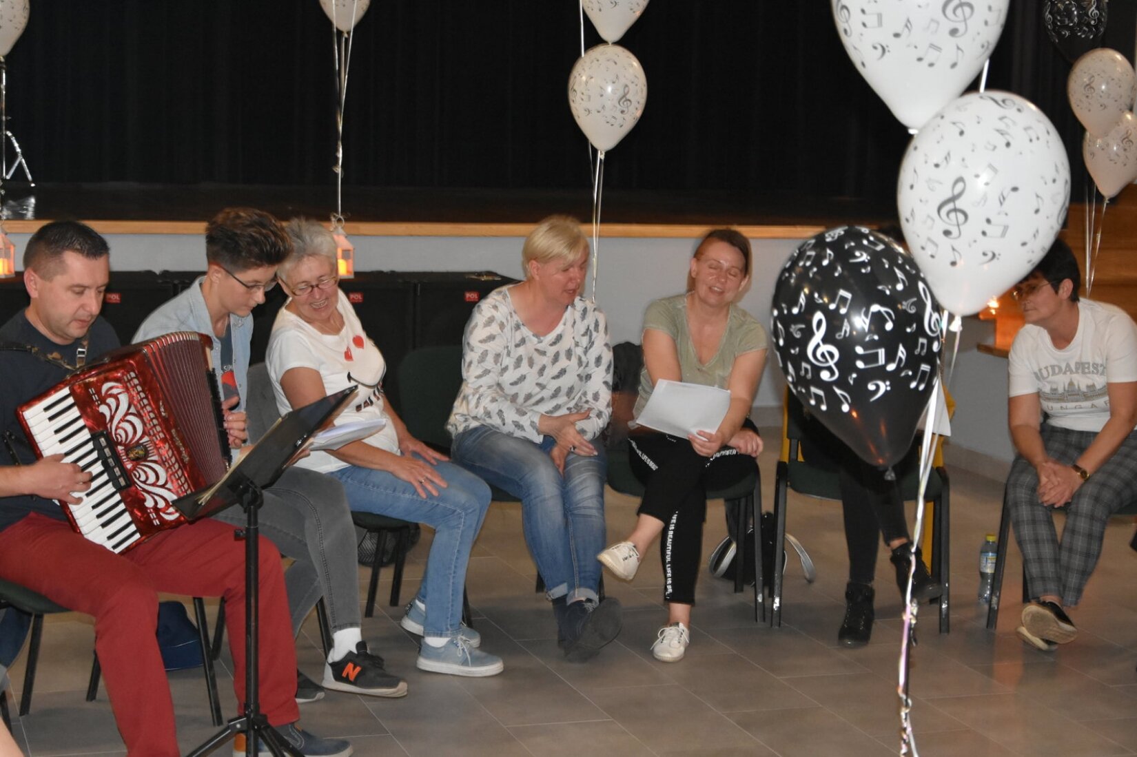 Zdjęcie przedstawia warsztaty w Gminnym Ośrodku Kultury w Wilczynie. Na zdjęciu znajduje się grupa ludzi siedząca na krzesłach. Mężczyzna po lewej stronie zdjęcia gra na akordeonie. Pozostałe osoby trzymają w rękach kartki oraz śpiewają. W tle zdjęcia znajdują się balony oraz scena.