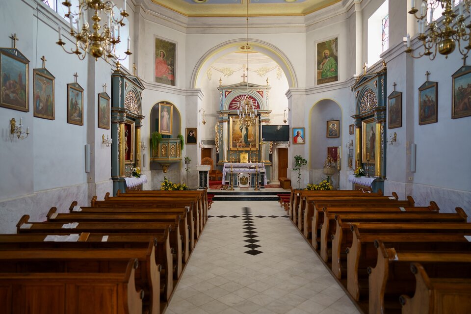 Zdjęcie przedstawia wnętrze kościoła nakierowane w stronę prezbiterium od strony chóru. Widoczny trójboczny układ prezbiterium z dwoma wejściami i dwoma bocznymi kaplicami w niszach figuralnych. Ściany podzielone są pilastrami i gzymsem, wyznaczającym świątyni dwie kondygnacje. W górnej części prezbiterium zamkniętej półkoliście wydzielone są po jej obu stronach nisze na obrazy. W wykuszach okiennych są witraże. Ściany zdobią klasycystyczne ołtarze. W znajdującym się w prezbiterium ołtarzu z wizerunkiem Wniebowziętej NMP znajduje się w półokrągłym tympanonie typowe dla klasycyzmu wyobrażenie „oka Opatrzności”. W niszy figuralnej po prawej stronie kościoła jest chrzcielnica. 