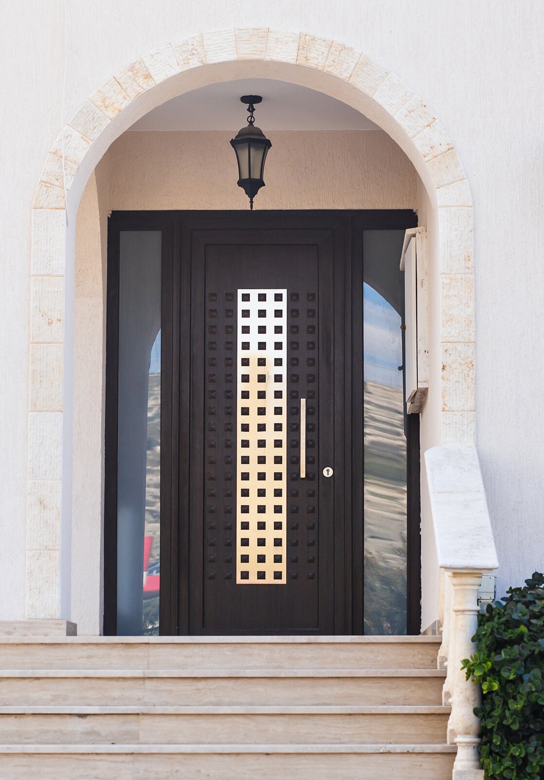 Zdjęcie przedstawia solidnie wykonane drewniane drzwi, na której powierzchni znajduje się kilkanaście rzędów wypukłych kwadratowych elementów udekorowanych dodatkowo metalowym pasem przechodzącym przez środek skrzydła. Po lewej i prawej stronie znajdują się wąskie przeszklone segmenty konstrukcji.