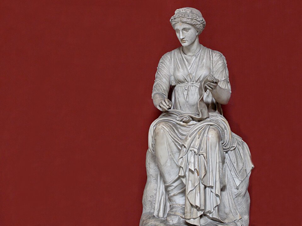 Zdjęcie przedstawia marmurowy posąg młodej kobiety. Kobieta ma pociągłą twarz i regularne rysy. Ubrana jest w długą, drapowaną szatę. Siedzi. W lewej ręce trzyma zwój papirusu lub pergaminu.