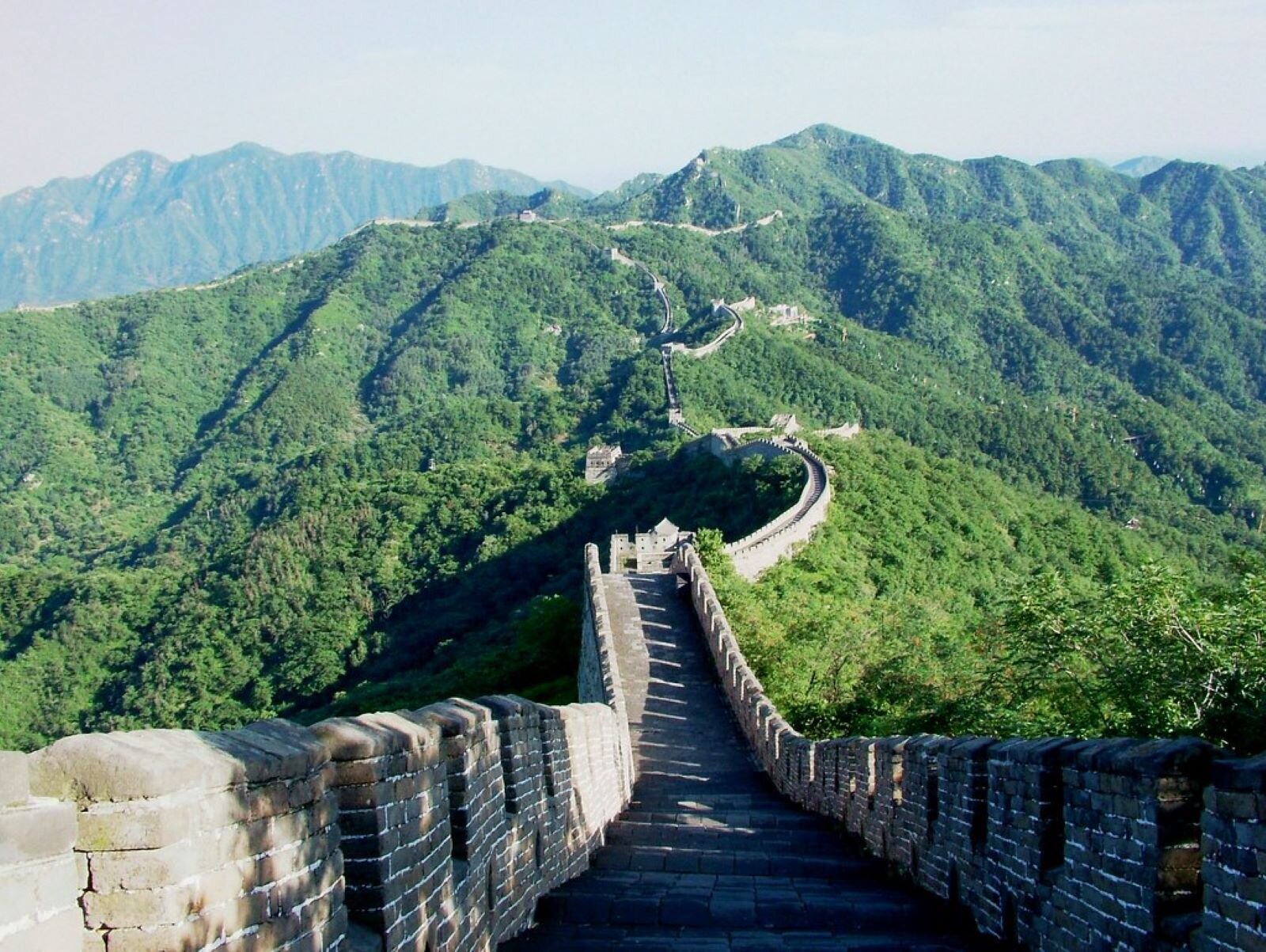 Zdjęcie przedstawia fragment Wielkiego Muru Chińskiego, najsławniejszej kamiennej budowli obronnej starożytnego państwa Chin. Mur spełniał funkcje obronne, ale i komunikacyjne. Fotografia ukazuje wysokie partie gór pokryte roślinnością i wnętrze muru widoczne z góry. Wstęga budowli wije się łamaną linią aż po horyzont.