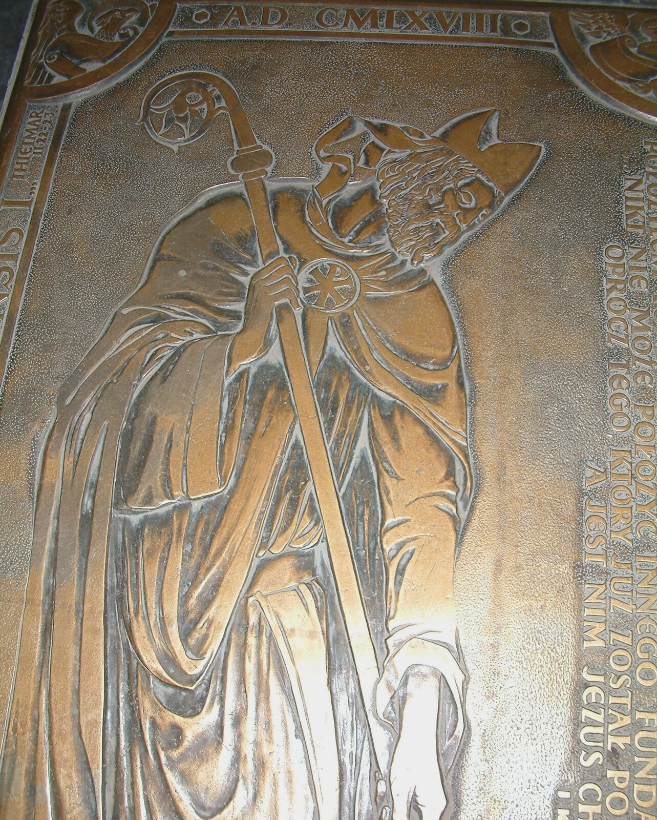 Ilustracja przedstawia płytę, na której widnieje wyryta postać duchownego ubranego w czapkę biskupią tak zwaną mitrę. Mitra ma formę wydłużonej czapki, składającej się z dwóch wysokich, połączonych ze sobą trójkątów, z opadającymi z tyłu dwiema wstążkami. Na plecach ma płaszcz, długi, zapinany pod szyją na klamrę w kształcie koła. W rękach trzyma pastorał, długa laskę o ślimakowatym zakończeniu. Na brzegach płyty znajdują się napisy.