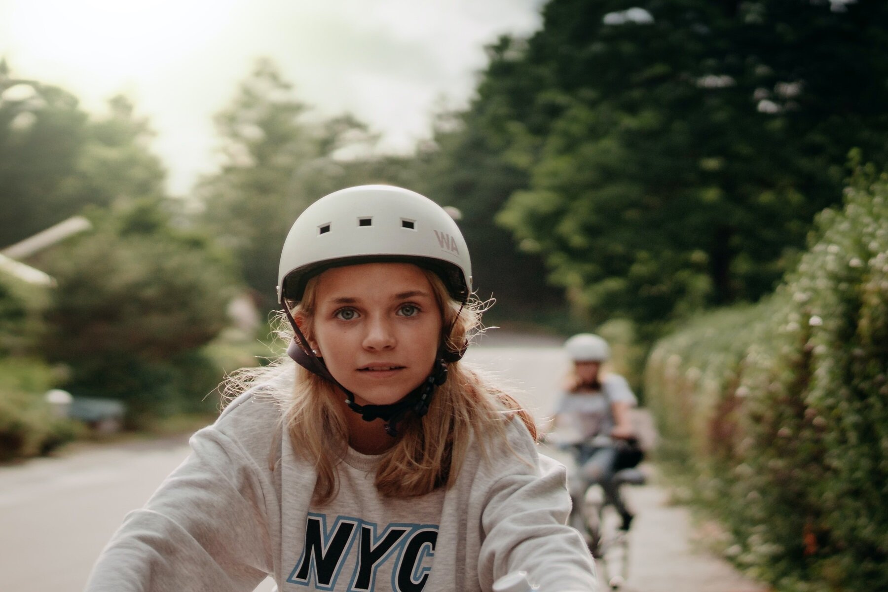 Zdjęcie przedstawia na pierwszym planie dziewczynkę od pasa w górę, jadącą na rowerze. Dziewczynka ma pogodną twarz i spojrzenie skierowane w obiektyw. Ma rozpuszczone, dłuższe blond włosy, wystające spod białego kasku ochronnego. Ubrana jest w szarą bluzę z literami NYC. Jej ramiona są wyciągnięte do przodu, więc prawdopodobnie ręce trzyma na kierownicy. W tle widoczna rozmyta postać jadąca na rowerze oraz droga i zielone drzewa i krzewy po obu jej stronach. 