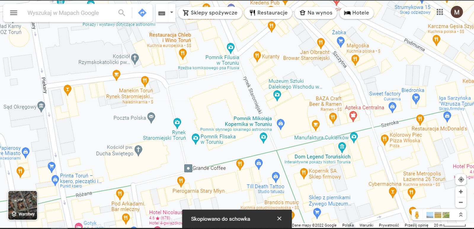 Zrzut ekranu prezentujący widok centrum Torunia w Google Maps. Ulice oznaczono białymi liniami, budynki szarymi lub jasnopomarańczowymi konturami. Podano nazwy ulic. Dodatkowo sygnaturami oznaczono ważniejsze punkty usługowe, zabytki i urzędy. U dołu ekranu wyświetla się czarne pole z napisem „Skopiowano do schowka”.