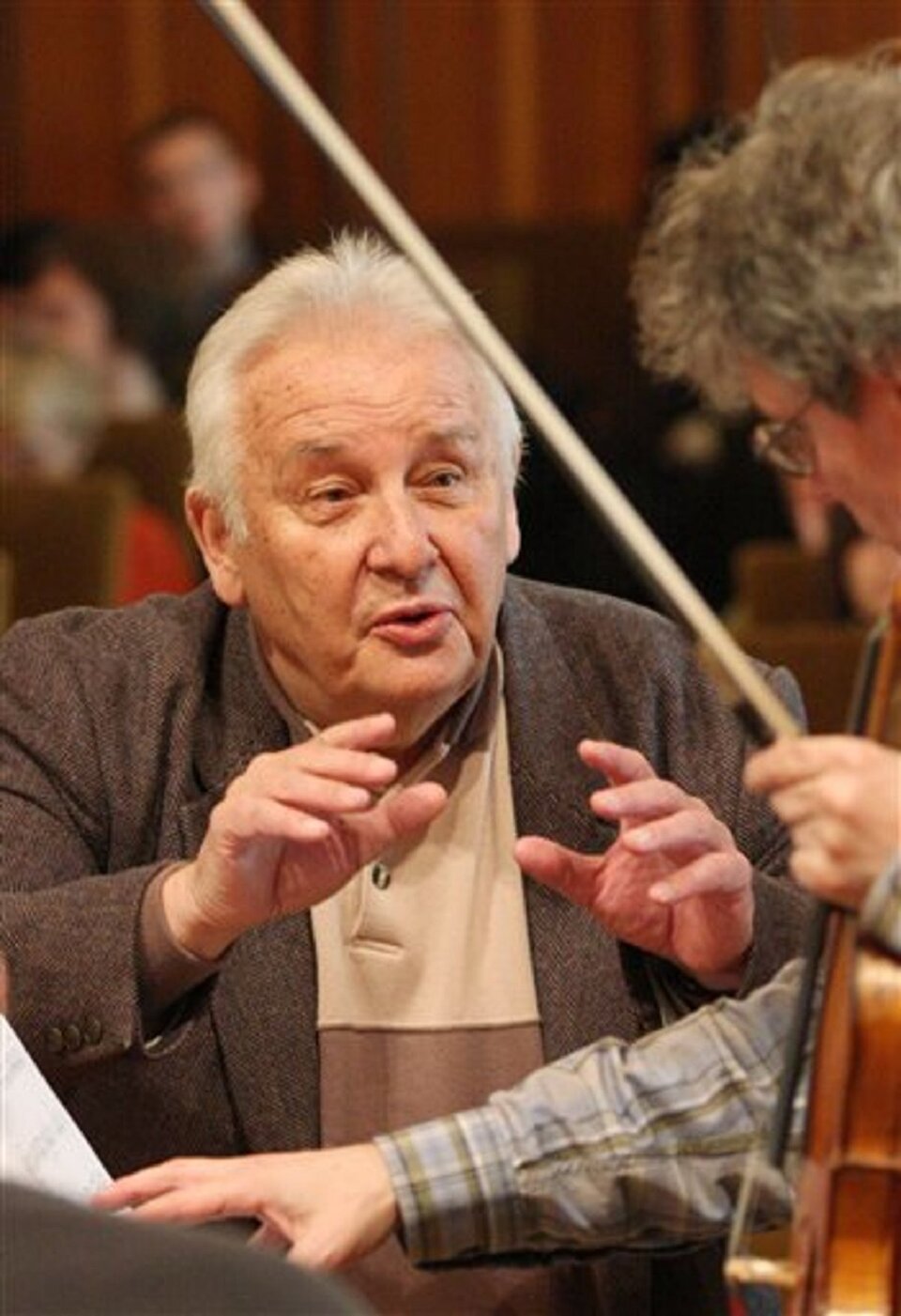 Fotografia przedstawia: Henryk Mikołaj Górecki podczas próby z zespołem Kronos Quartet. Na zdjęciu znajduje się siwy kompozytor, rozmawiający z drugą osobą. W prawym rogu widoczny jest fragment skrzypiec ze smyczkiem.
