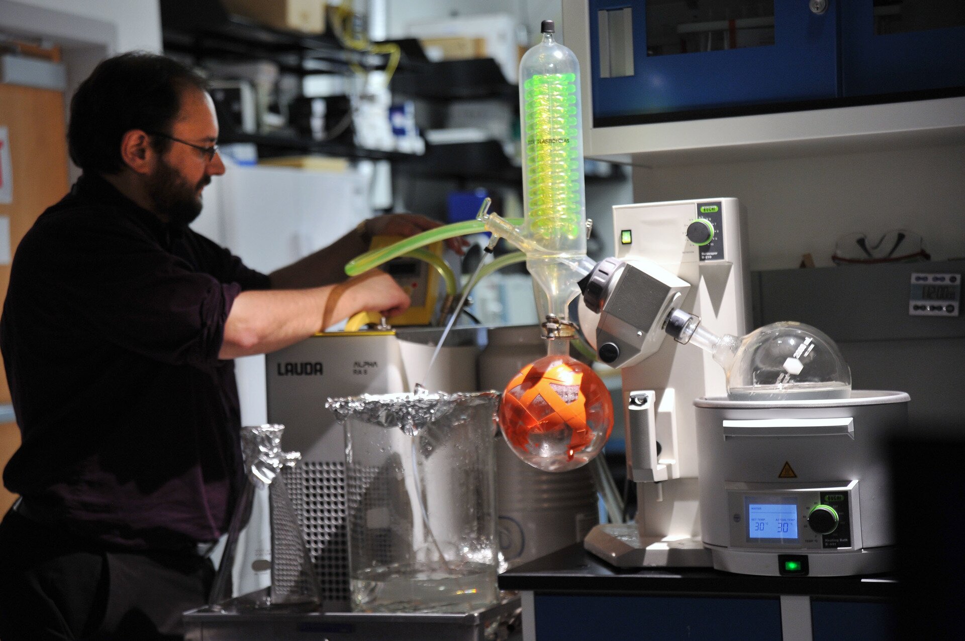 Ilustracja przedstawia mężczyznę w laboratorium chemicznym. Mężczyzna ma brodę, okulary nie jest ubrany w strój ochronny. Obsługuje specjalistyczny sprzęt laboratoryjny.