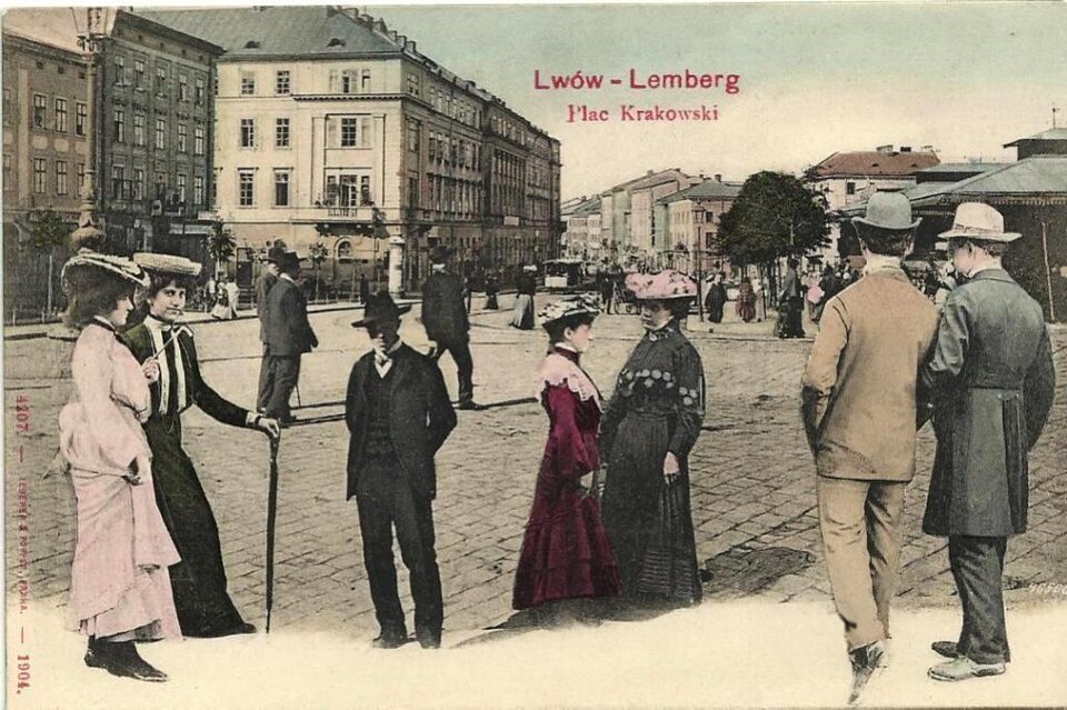 Na fotografii widać dawny Lwów, Plac Krakowski. Przedstawiona jest scena uliczna z początku XX wieku – na pierwszym planie elegancko ubrani przechodnie, kobiety w długich sukniach i ozdobnych kapeluszach oraz mężczyźni w garniturach, płaszczach i kapeluszach. W tle widać zabudowę miejską z kamienicami i szyldami, brukowany plac oraz dalszy ruch pieszy.