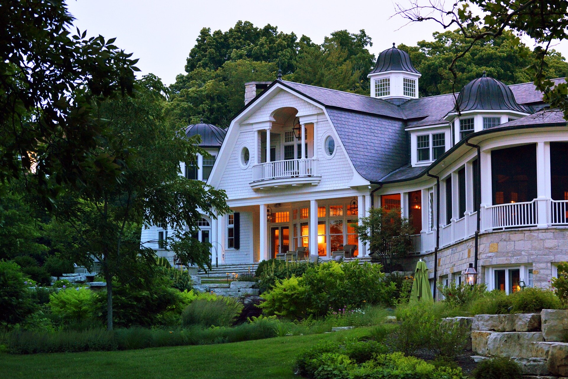 Zdjęcie pierwsze przedstawia elegancki, duży, murowany dom stojący na niewielkim wzniesieniu. Prowadza do niego szerokie schody. W centralnej części znajduje się wsparty na kolumnach ganek. Jest też duży, zadaszony taras. Przed domem znajduje się rozległy trawnik z licznymi krzewami i drzewami. 