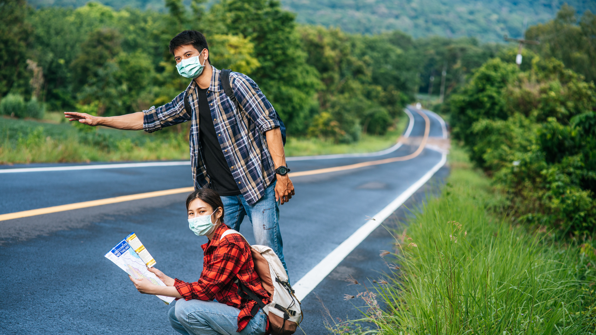 Zdjęcie przedstawia dziewczynę i chłopaka, którzy znajdują się przy drodze. Dziewczyna kuca, w rękach trzyma mapę. Chłopak stoi przy drodze i wyciąga jedną rękę w celu zatrzymania jakiegoś samochodu. Oboje ubrani są w koszule w kratę oraz dżinsy. Na twarzach mają maseczki ochronne. W tle widoczna jest pusta droga i drzewa po jednej i po drugiej stronie.