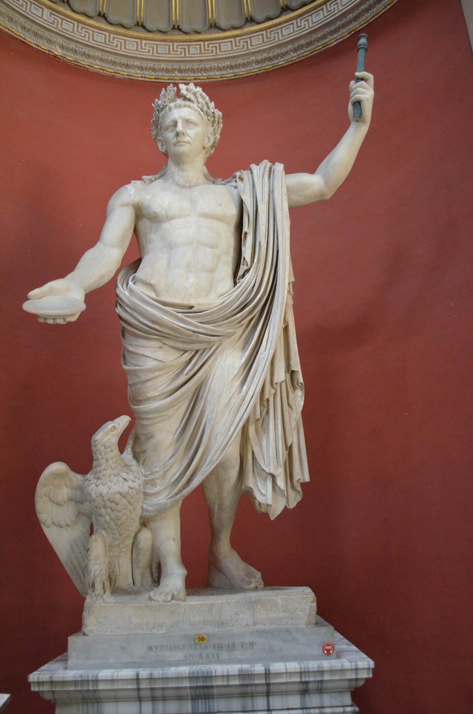 Ilustracja o kształcie pionowego prostokąta przedstawia rzeźbę cesarza Klaudiusza. W swojej lewej ręce, uniesionej do góry dzieży berło. W prawej trzyma naczynie. Obok jego prawej nogi stoi orzeł. Na głowie ma wieniec z liści laurowych. Ciało postaci odziane jest od połowy w materiał, zawieszony na lewym ramieniu. Klaudiusz stoi w kontrapoście, z głową lekko uniesioną do góry, jest poważny. Ukazany został na bordowym tle. 