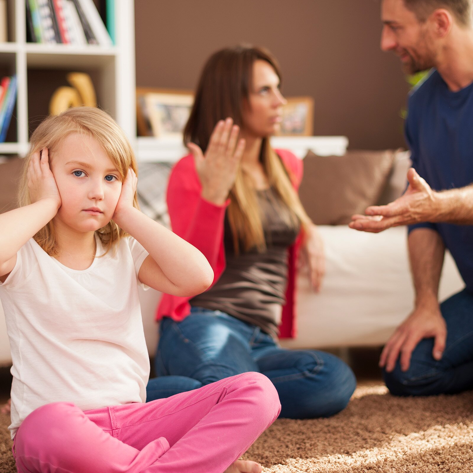 Ilustracja przedstawia kobietę i mężczyznę siedzących na podłodze pochłoniętych dyskusją i gestykulujących. Z boku po turecku siedzi odwrócona do nich plecami mała dziewczynka w wieku szkolnym i zasłania uszy dłońmi, jakby nie chciała słyszeć kłótni.