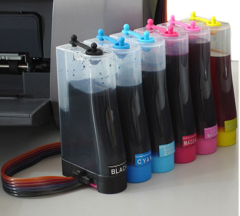 Zdjęcie przedstawia zbiorniki do tuszy. Widoczne jest 6 plastikowych wysokich zbiorników, ułożonych szeregowo, wypełnionych czarną substancją – tuszem, które mają podstawy w różnych kolorach od lewej: czarny, ciemnoniebieski, niebieski, czerwony, różowy, żółty, oraz odpowiadające im kolorystycznie zatyczki otworów w górnej części zbiorników. W tle widoczna część drukarki.