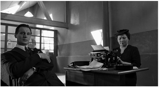 Zdjęcie przedstawia wnętrze fabryki. Robotnicy malują ściany. W sali stoi biurko, przy którym siedzi przy maszynie do pisania dojrzała kobieta w niewielkim kapeluszu i z papierosem w ustach. Naprzeciw niej siedzi lewym bokiem do kobiety Schindler. Patrzy się przed siebie.