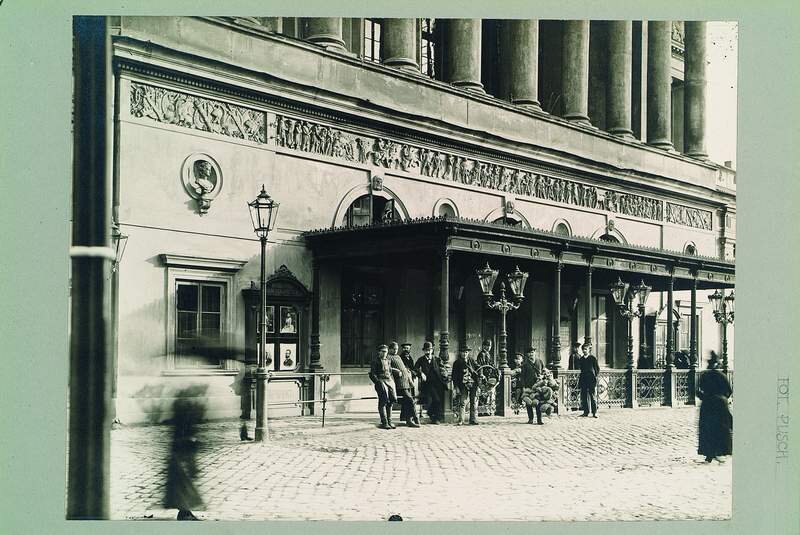 Historyczna, czarno-biała fotografia przedstawiająca elegancki budynek z klasycystyczną fasadą, zdobioną fryzami i kolumnami. Przed wejściem znajduje się zadaszony ganek z ozdobnymi latarniami. Na brukowanej ulicy widać grupę mężczyzn siedzących na ławce, kilku stojących oraz przechodniów – niektórzy są rozmazani, co sugeruje ruch w trakcie wykonywania zdjęcia.