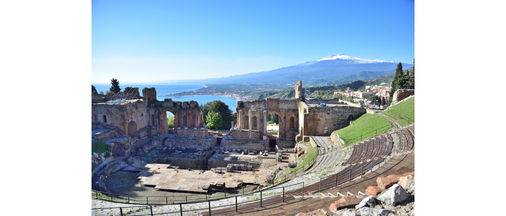 Zdjęcie na pierwszym planie przedstawia teatr grecki w sycylijskim miasteczku Taormina. W tle widoczna jest zatoka, nad którą leży Taormina oraz wulkan Etna.