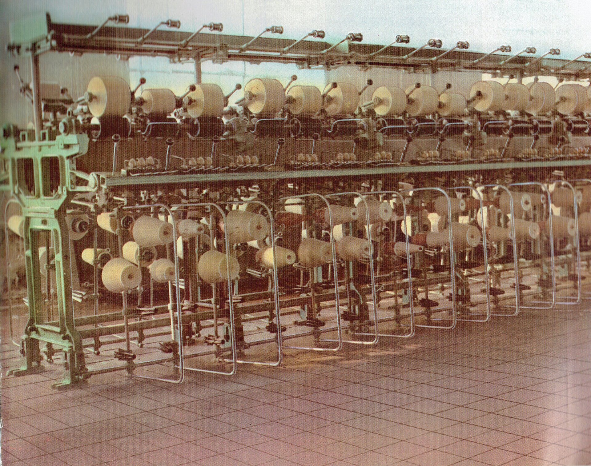 Zdjęcie przedstawia łączniarkę. Na maszynie zamontowane są w rzędach szpulki z nićmi w górnej i w dolnej części maszyny. Widoczne są nitki przechodzące przez wałki i inne elementy mechanizmu maszyny. 