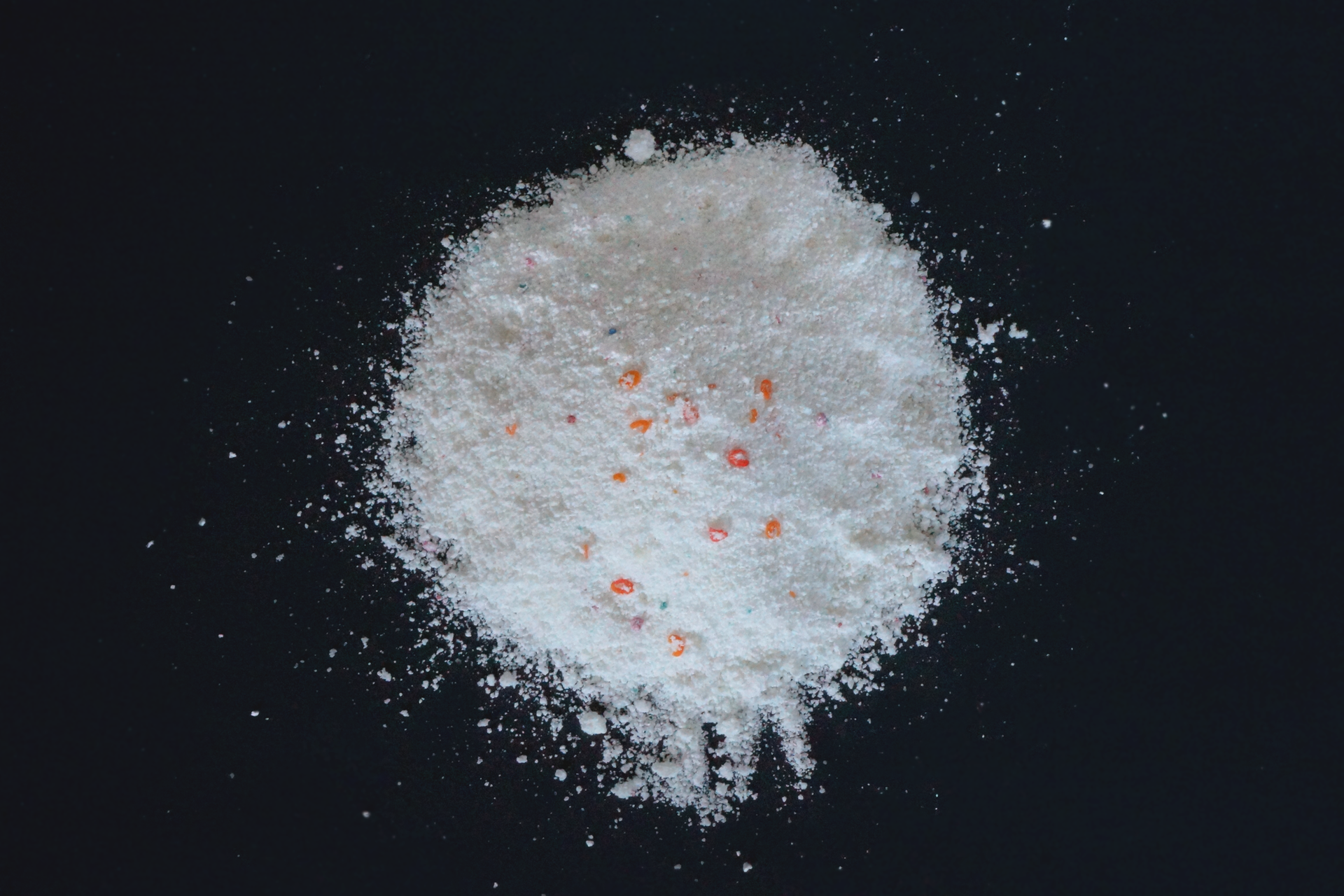 Fotografia przedstawia biały proszek do prania z pomarańczowymi granulkami.