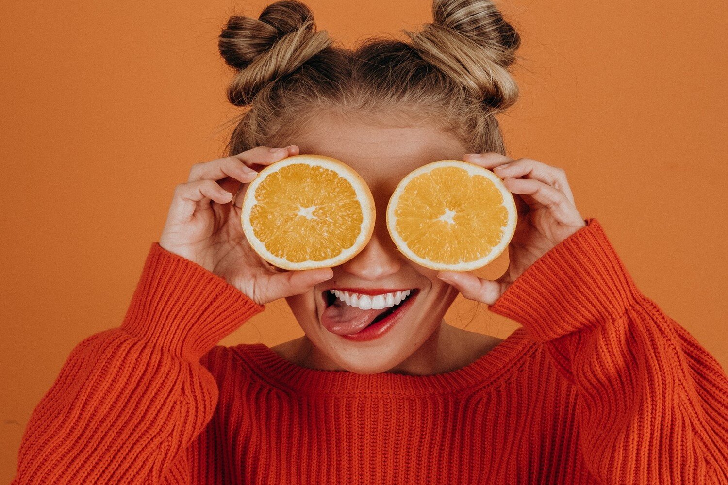 Zdjęcie przedstawia młodą kobietę od klatki piersiowej w górę. Kobieta trzyma w dłoniach krążki pomarańczy, zasłaniając sobie nimi oczy. Na twarzy kobiety jest szeroki uśmiech, odsłaniający górne zęby spod których kobieta wstawiła język skierowany do boku. 