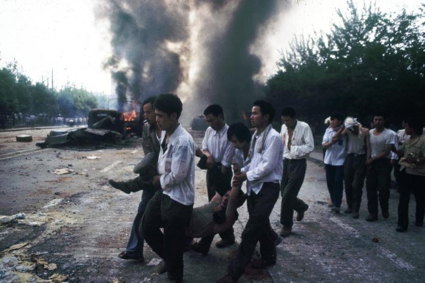 Zdjęcie przedstawia ulicę, na której kilku mężczyzn niesie za nogi i ręce kobietę. Najprawdopodobniej jest ranna. Za nimi dwóch mężczyzn podtrzymuje mężczyznę rannego w głowę, którą ma zawiniętą w koszulę. W tle palące się samochody, żywy ogień i czarny dym. 