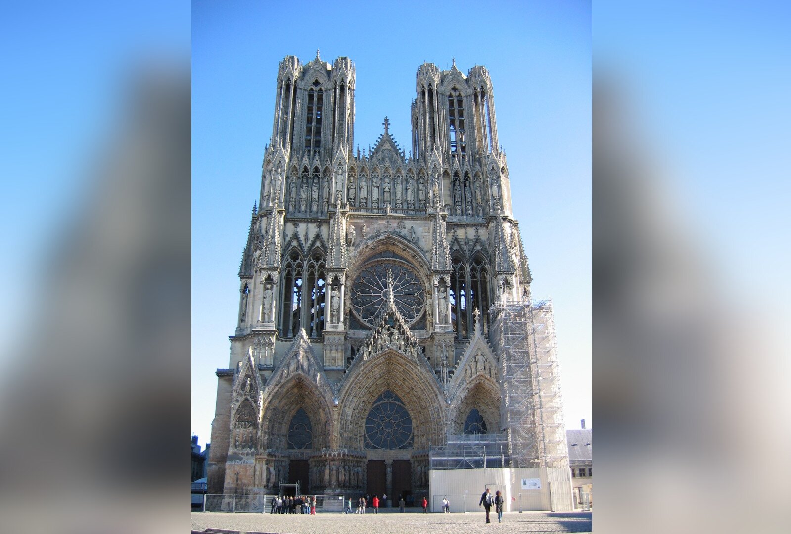 Zdjęcie przedstawia front gotyckiej katedry. Po bokach składa się z dwóch wież, pośrodku ma duże, okrągłe okno. Do świątyni prowadzą wysokie drzwi umieszczone w strzelistym łuku. Po prawej stronie budynku stoją rusztowania. Przed katedrą jest plac, po którym spacerują ludzie.