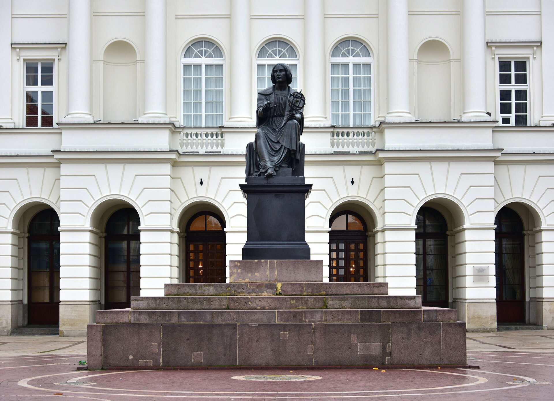 Na zdjęciu pomnik w kolorze ciemnoszarym. Przedstawia siedzącego mężczyznę. Ma proste włosy średniej długości. Ubrany jest w szatę do kostek. Prawą rękę ma wyciągniętą do przodu, a w lewej trzyma astrolabium. Pomnik stoi na ciemnoszarym podeście, dookoła którego są czerwono - szare schody. Przed pomnikiem jest bordowy plac z beżowymi okręgami. W tle znajduje się jasny budynek z łukowymi oknami. Okna na parterze są w kolorze czarnym, a na piętrze w białym. 