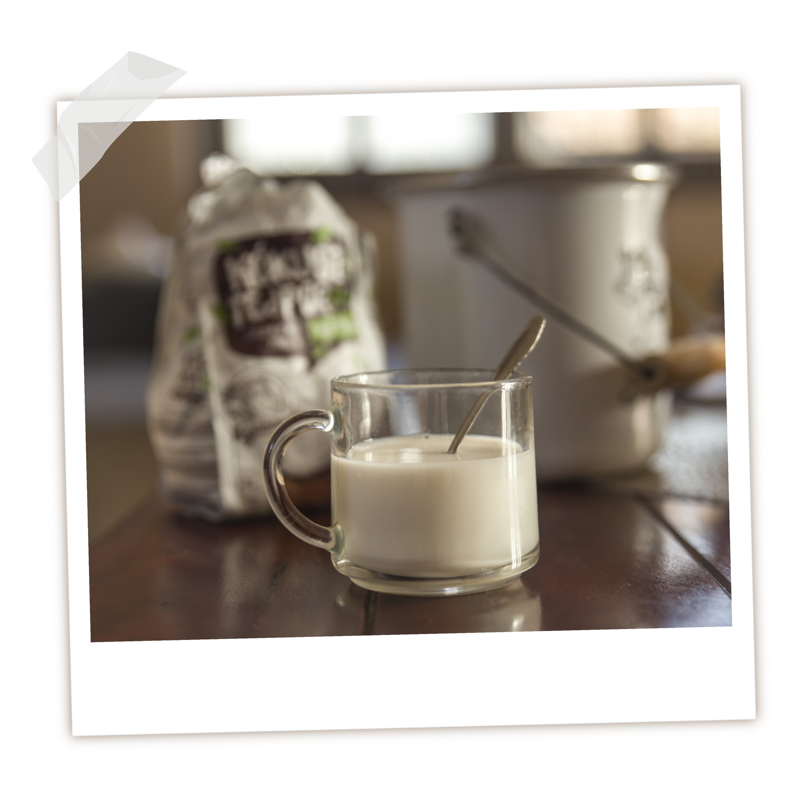  Na zdjęciu widoczny jest jogurt sojowy w szklance. Szklanka leży na blacie. W tle widoczna jest zapakowana kawa i garnek. 