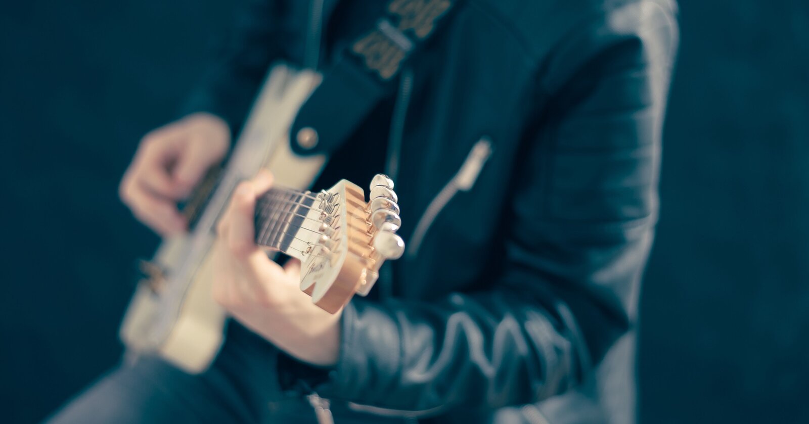 Fotografia przedstawia osobę grającą na gitarze. Pośrodku, na pierwszy planie, widoczny jest sam gryf gitarowy. Reszta gitary oraz osoba ją trzymająca jest rozmyta. Osoba ma na sobie czarną skórzaną kurtkę. Widać jedynie jej ręce oraz tułów. Tło jest jednolicie czarne.